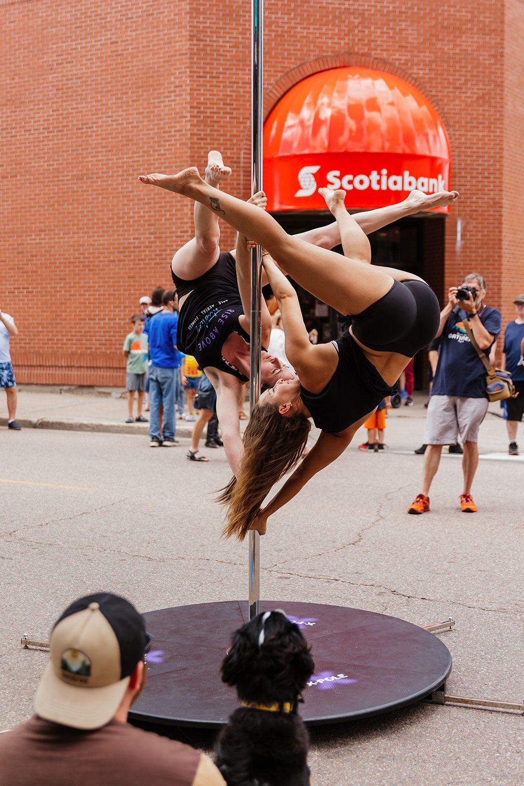 An aerialist inverted on a vertical pole in front of a street crowd.