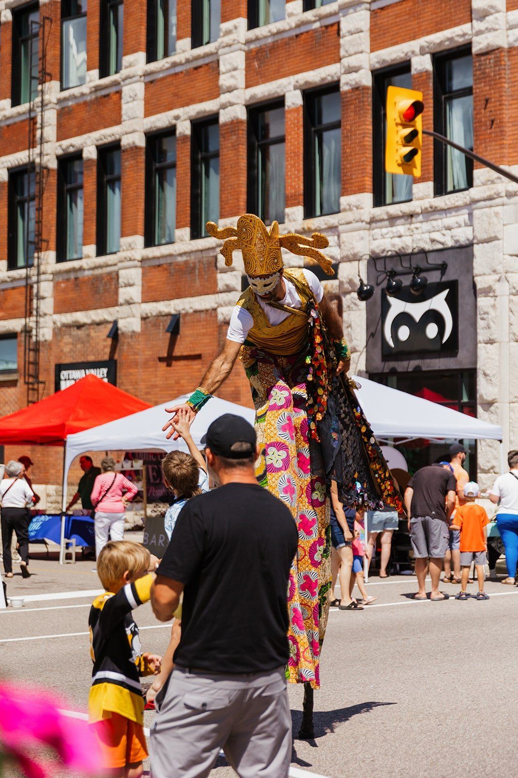 A towering giraffe puppet leaning down to meet a child at the roadside.