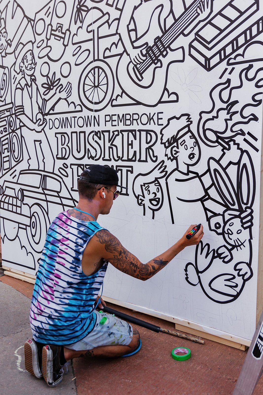 An artist inking a large black-and-white Downtown Pembroke Buskerfest mural board.
