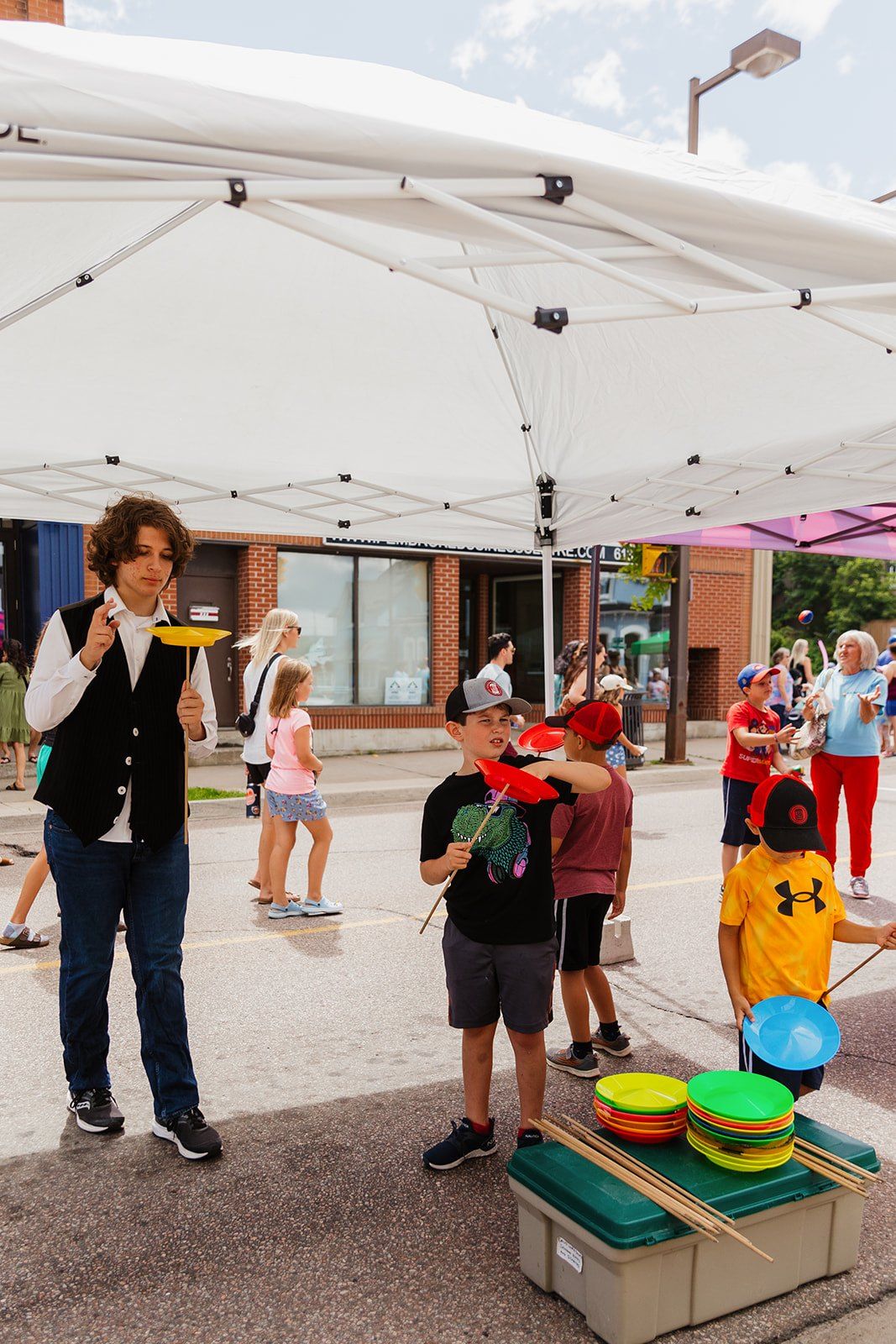 A performer teaching children to spin plates under a white tent on the street.