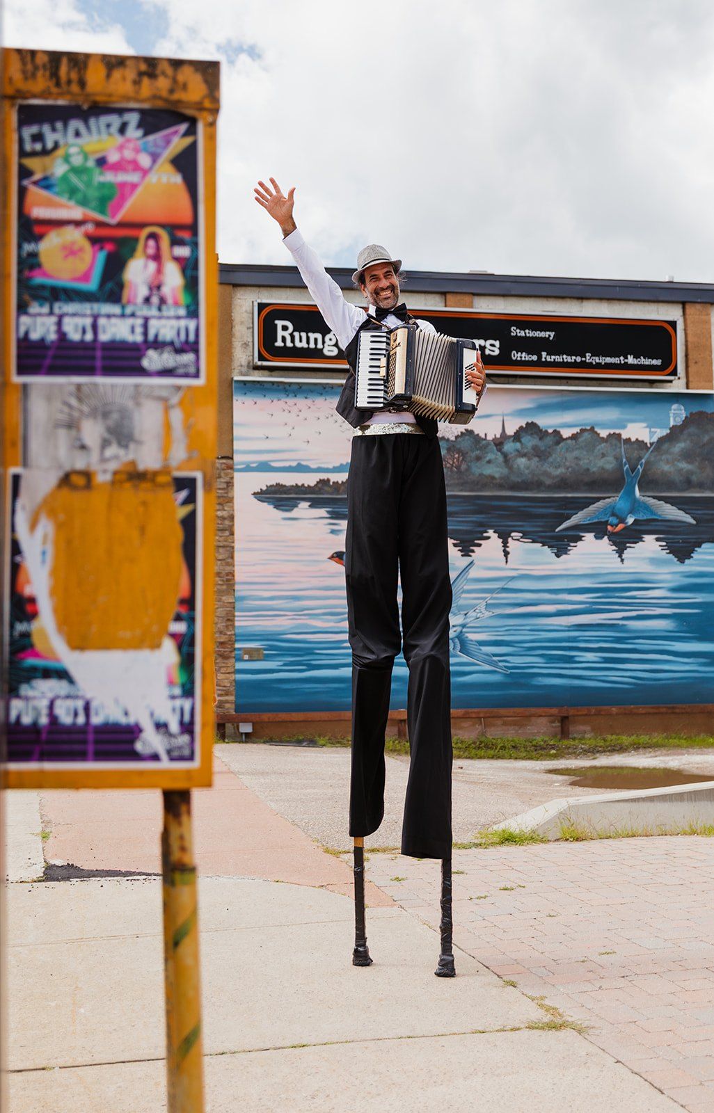 A stilt-walker playing an accordion beside a painted wall mural.