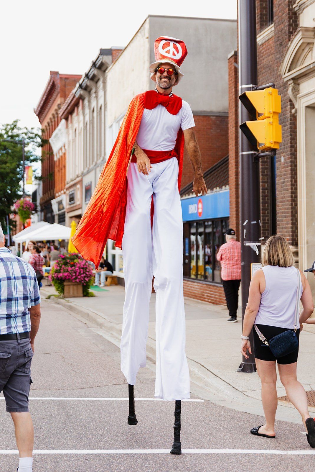 A stilt-walker in a red and white costume with a flowing cape, walking the centre of the street.